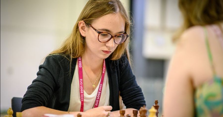 Alicja Śliwicka - podwójna mistrzyni Europejskich Igrzysk Akademickich Zdjęcie portretowe kobiety w okularach. Na szyi ma różową smycz, jej wzrok skierowany jest na szachownicę, która stoi przed nią.