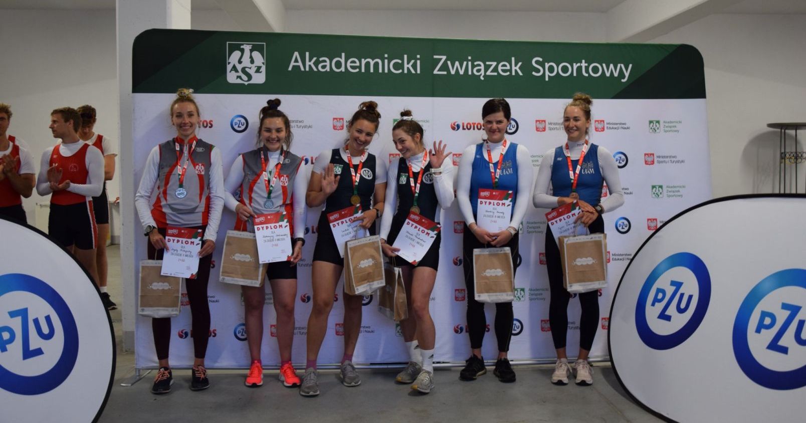 Po lewej stronie stoją Katarzyna Malinowska i Agata Zielepuha, srebrne medalistki w konkurencji dwójek podwójnych Sześć osób pozuje przed ścianką z napisem "Akademicki Związek Sportowy"