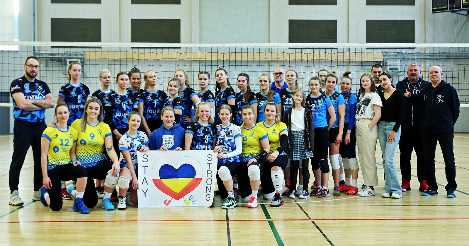  Grupa zawodniczek-siatkarek i trenerów stoi w hali sportowej przed siatką z plakatem o treści "stay strong" i sercem w  kolorach flagi Ukrainy