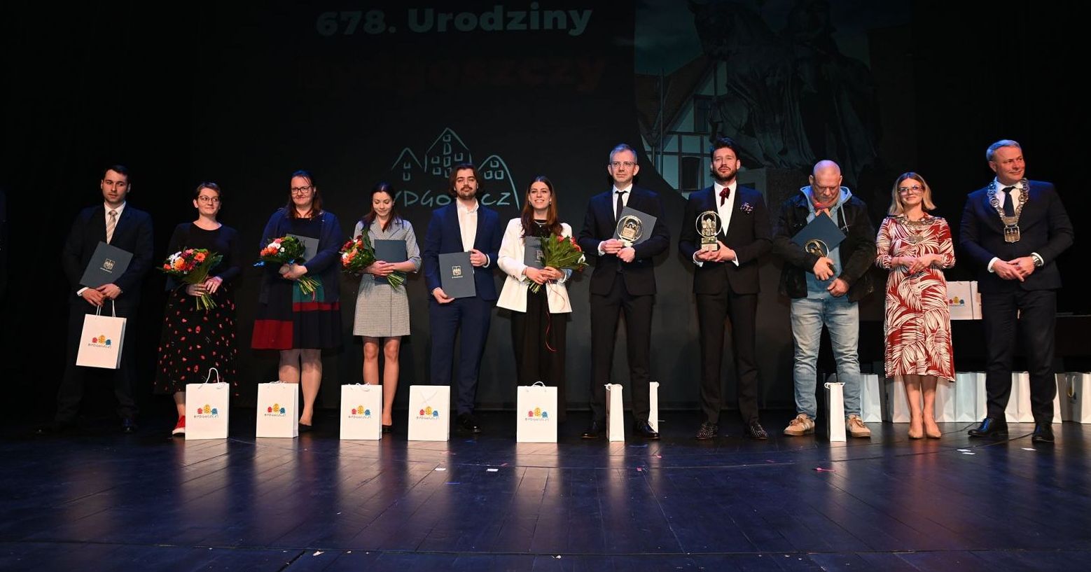 19 kwietnia Bydgoszcz świętowała swoje 678. urodziny, co było okazją do przyznania odznaczeń i nagród prezydenta miasta Nagrodzeni stoją w rzędzie na scenie i trzymają dyplomy, kwiaty lub statuetki.